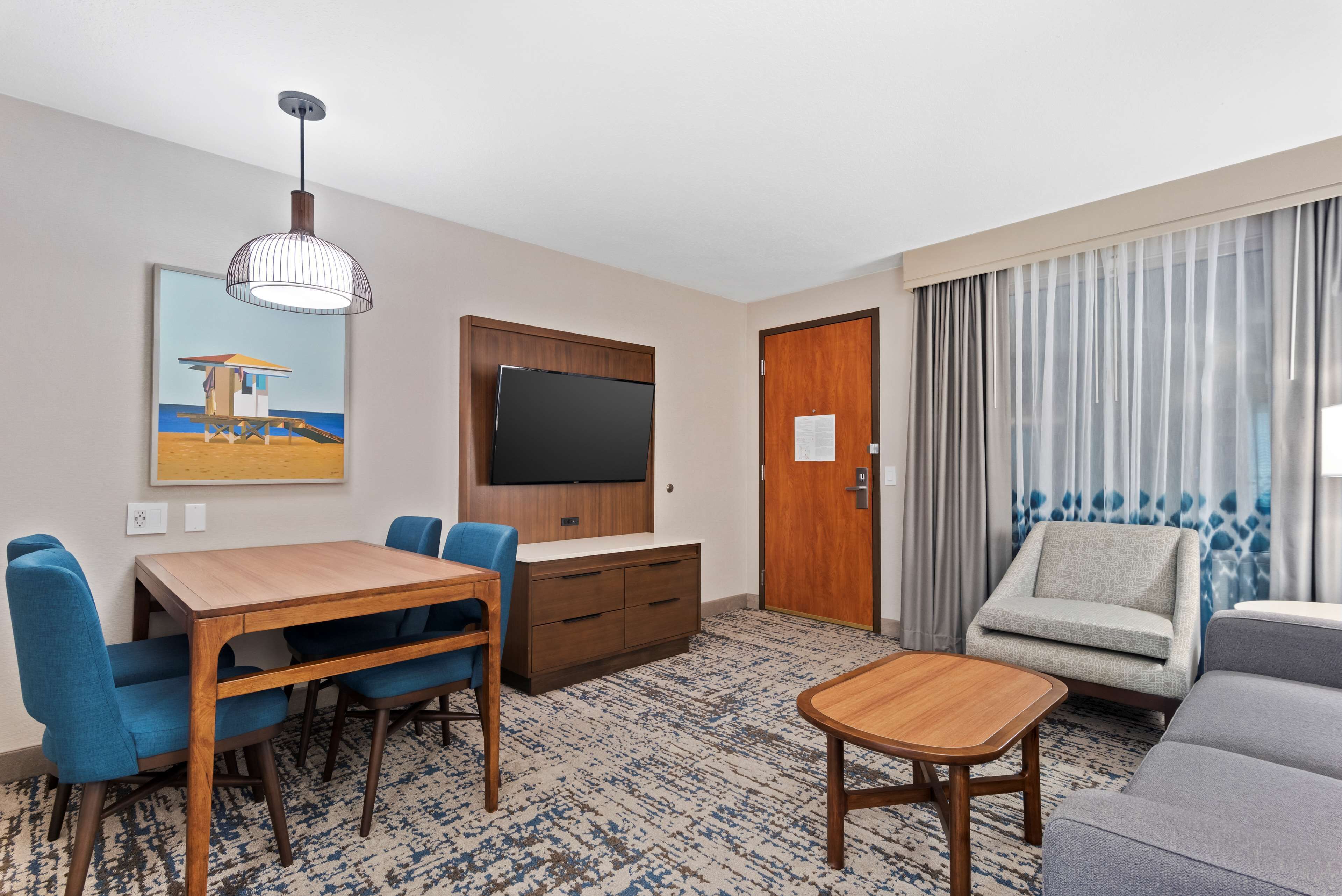 Habitación de hotel con una mesa de comedor de madera, sillas azules, un sofá gris, una mesita de noche de madera, un televisor de pantalla plana montado en la pared y un cuadro con una escena de playa.