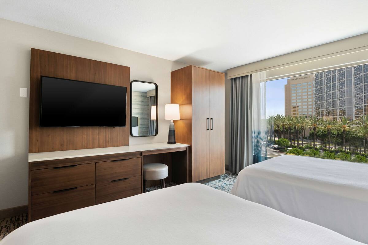 A hotel room with a large flat-screen TV, a desk with a lamp, a wardrobe, and two beds.