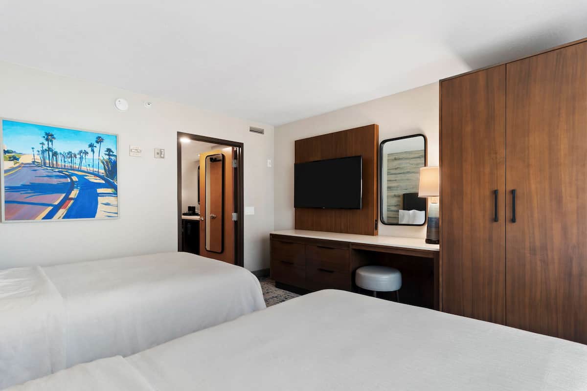 A hotel room with two beds, a TV, a desk with a mirror, and a painting of a road with palm trees.
