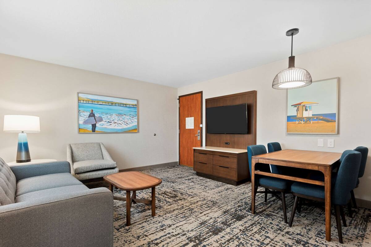 A hotel room with a sofa, armchair, small table, TV, and a dining area with chairs and a table.