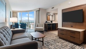 Une chambre d’hôtel équipée d’un canapé, d’une petite table, d’une télévision, d’une kitchenette et d’une grande fenêtre donnant sur la ville.
