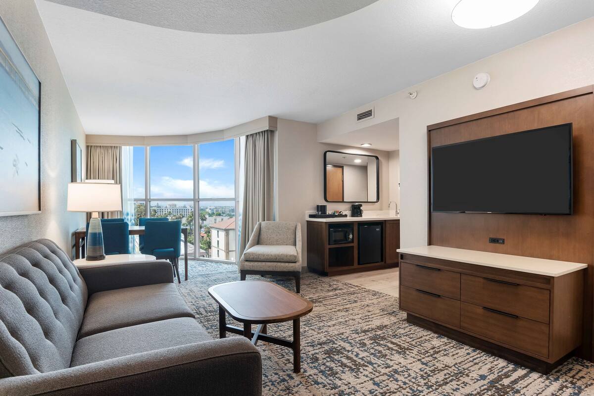 A hotel room with a sofa, a small table, a TV, a kitchenette, and a large window offering a city view.