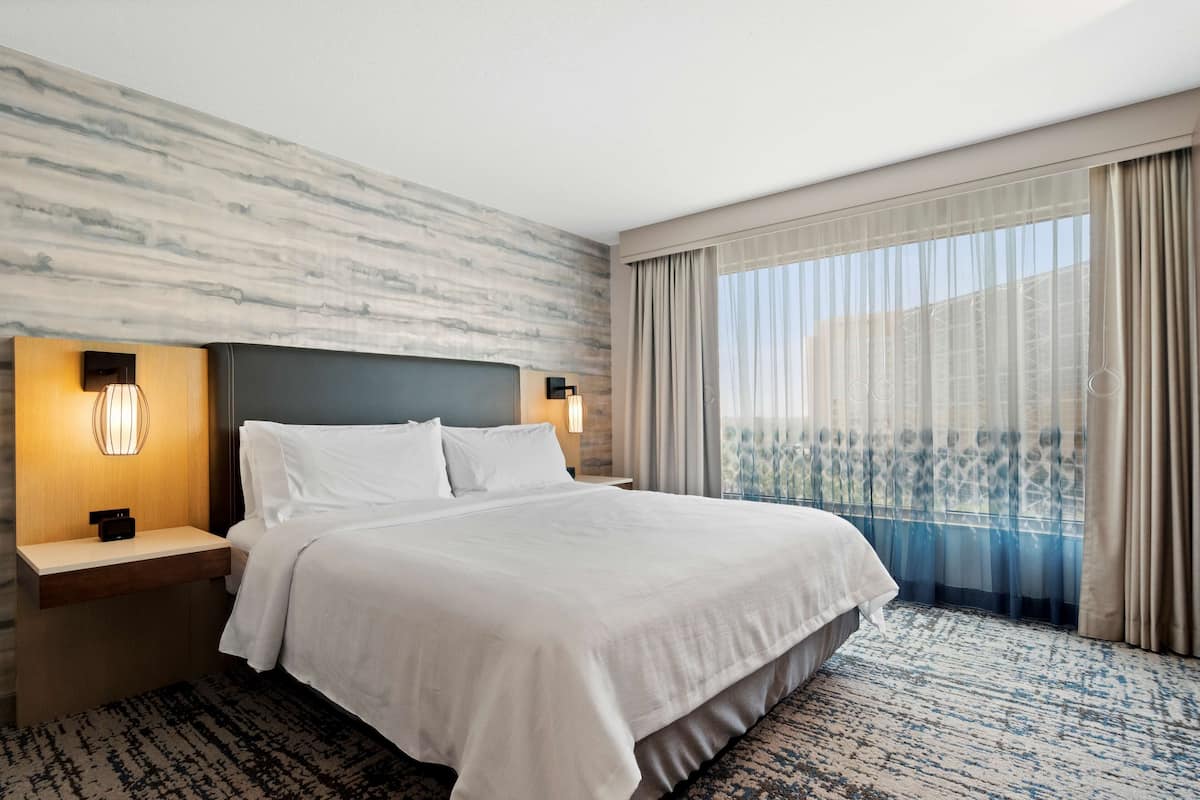 A hotel room with a large bed, two bedside tables with lamps, a marble-patterned wall, and a view of a balcony through sheer curtains.