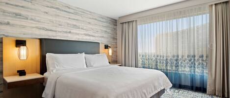 Habitación de hotel con una cama grande, dos mesitas de noche con lámparas, un muro con diseño de mármol y vistas a un balcón a través de cortinas de encaje.