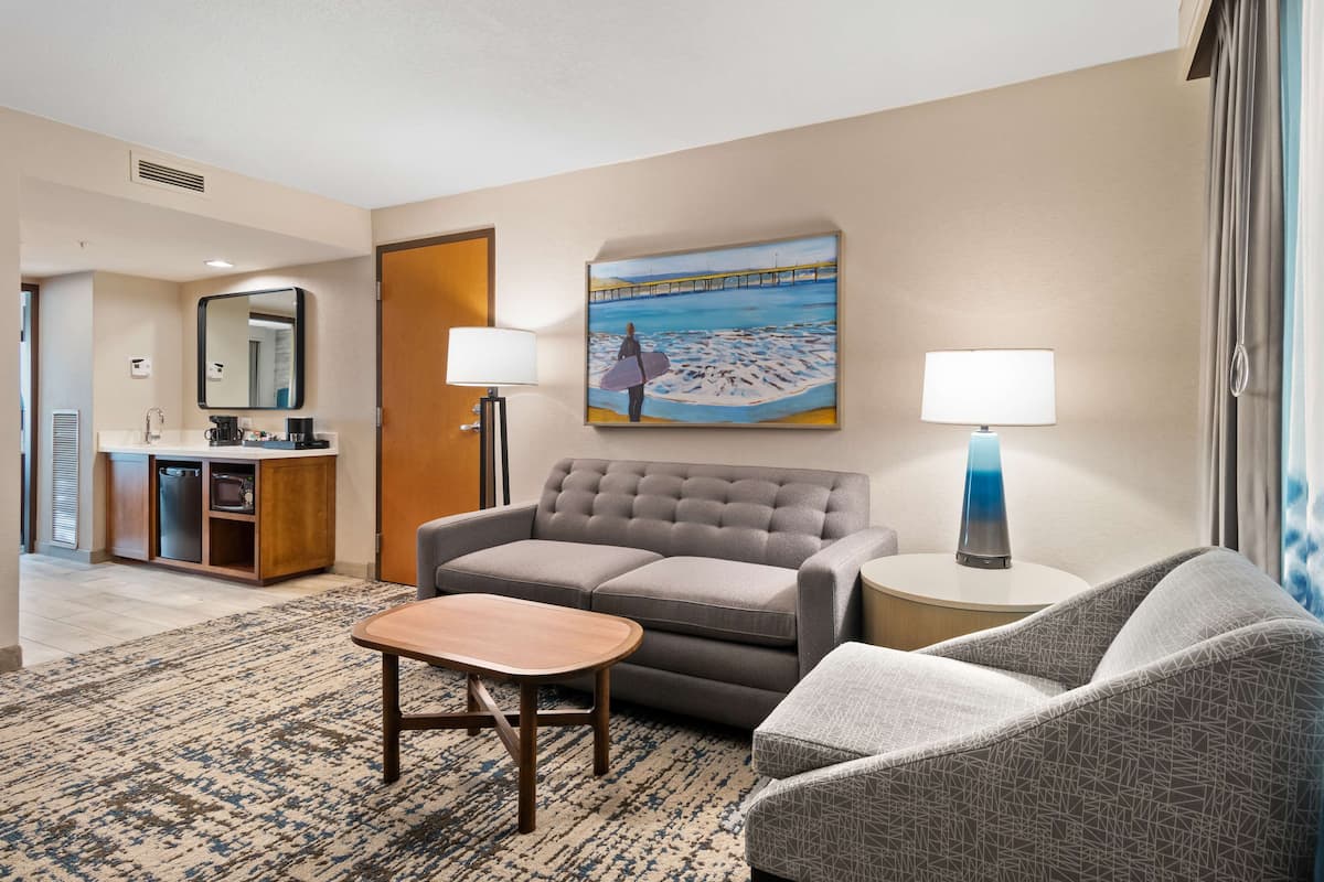 A hotel room with a grey sofa, a small table, a lamp, a painting of a pier, and a kitchenette.