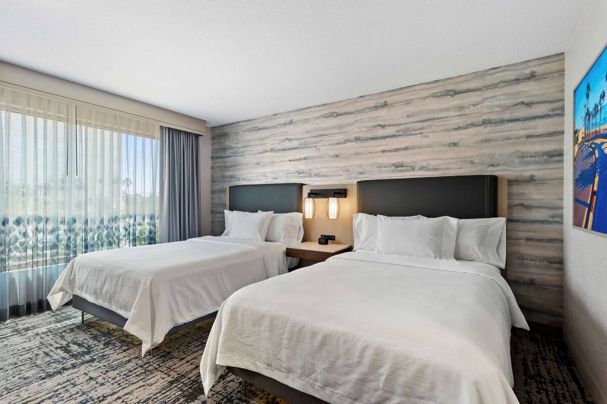 A hotel room with two beds, a large window with sheer curtains, a marble-patterned wall, and a painting on the right.