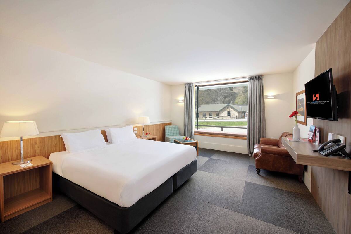 Ein Hotelzimmer mit einem großen Bett, einem Schreibtisch, einem Stuhl, einem Fernseher und einem Fenster mit Blick auf ein Gebäude.