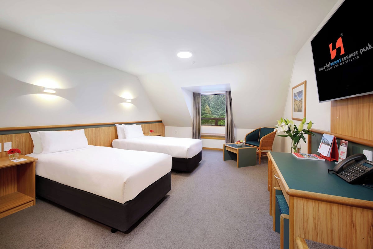 Ein Hotelzimmer mit zwei Betten, einem Schreibtisch mit Telefon, einem Sessel, einem kleinen Tisch mit Blumenvase und einem Fenster mit Vorhängen.
