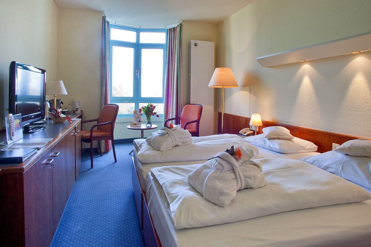 A hotel room with a bed, a desk with a TV, a chair, a lamp, and a window with curtains.