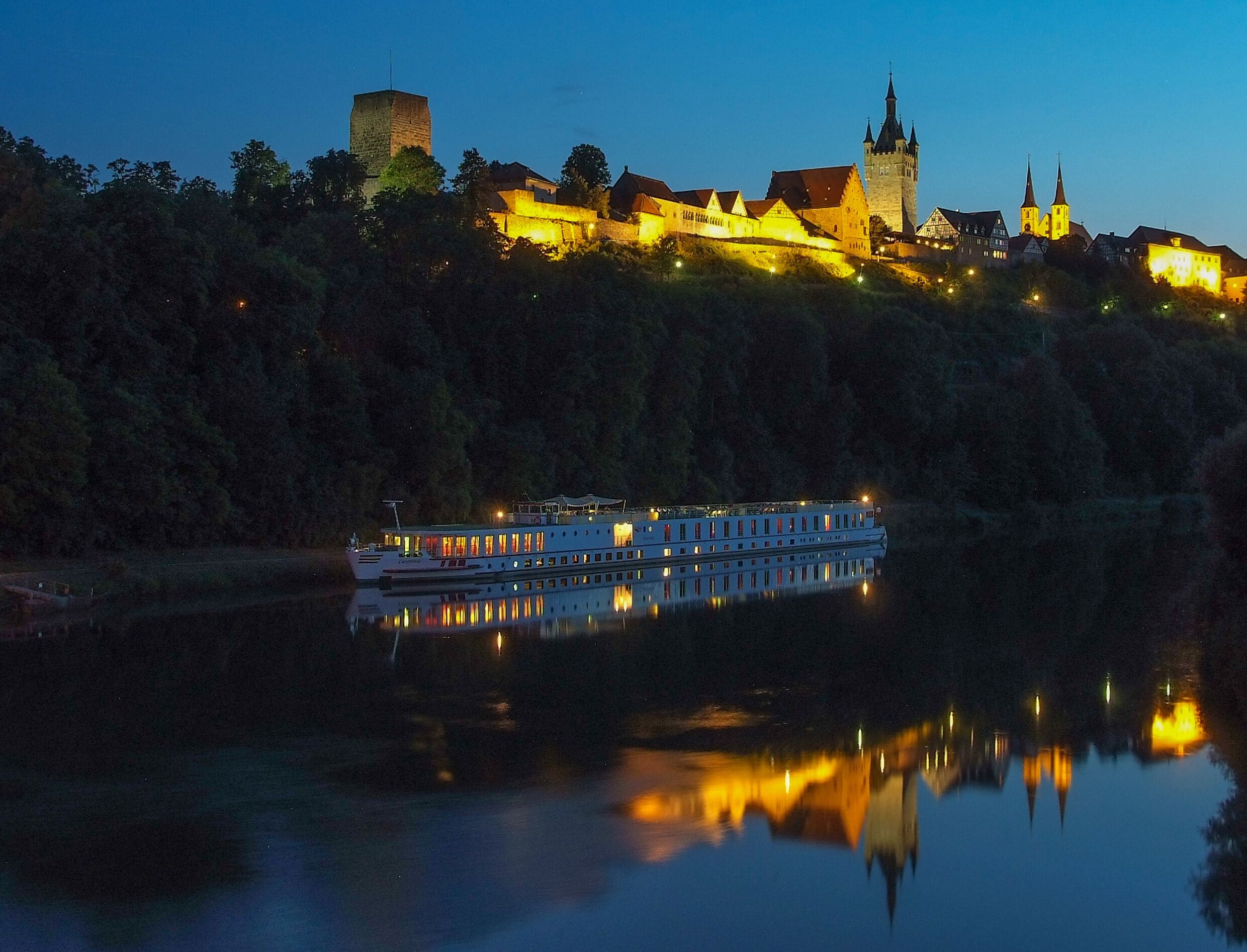 Ein Flusskreuzfahrtschiff liegt an einem Flussufer in einer Stadt mit historischen Gebäuden und einer Burg im Hintergrund.