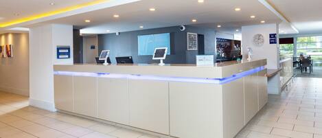 Zona de recepción de un hotel con diseño moderno, que incluye un mostrador de recepción, quioscos de registro digital y un reloj a la vista.