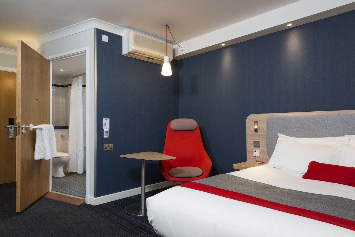 A hotel room with a bed, a red chair, a small table, a shower area, and an air conditioning unit.