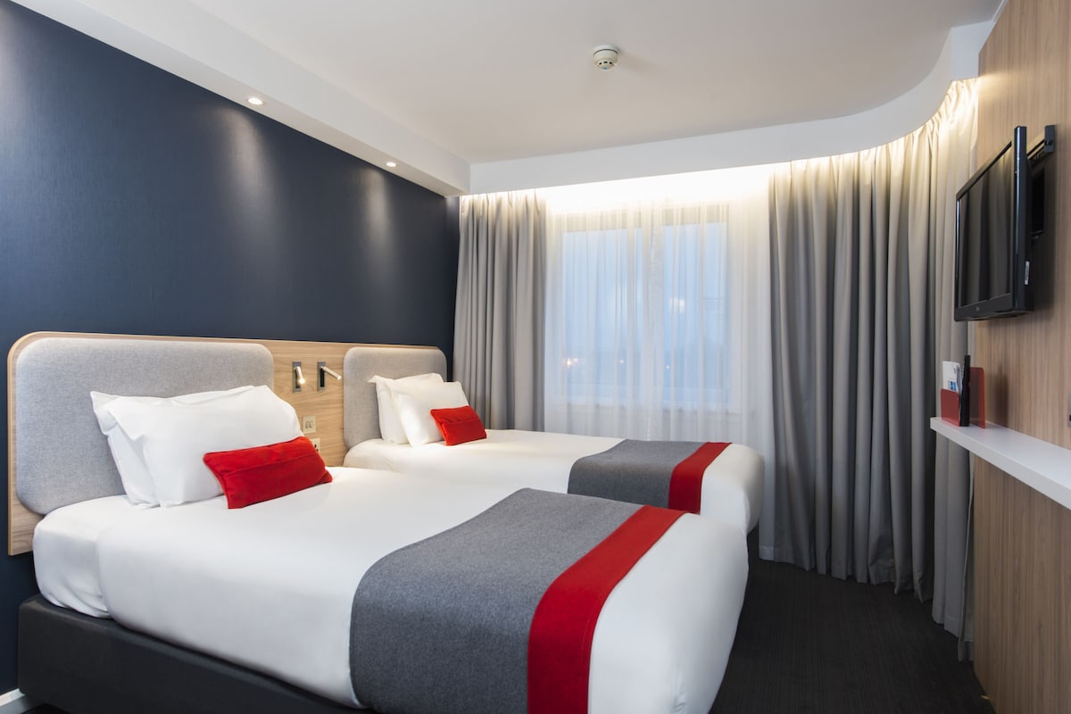 A hotel room with two beds, a flat-screen TV, a wooden headboard, and a large window with curtains.