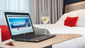 A laptop on a wooden table with a glass of white wine beside it, in a hotel room with a bed and red pillows. - Holiday Inn Express Milton Keynes by IHG (Milton Keynes)