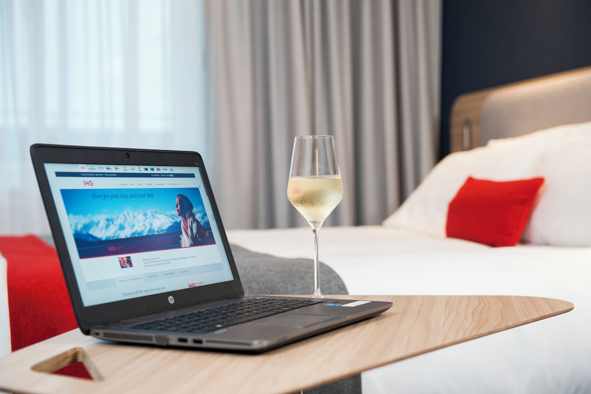 Una laptop sobre una mesa de madera, con una copa de vino blanco a su lado, en una habitación de hotel con cama y almohadas rojas.