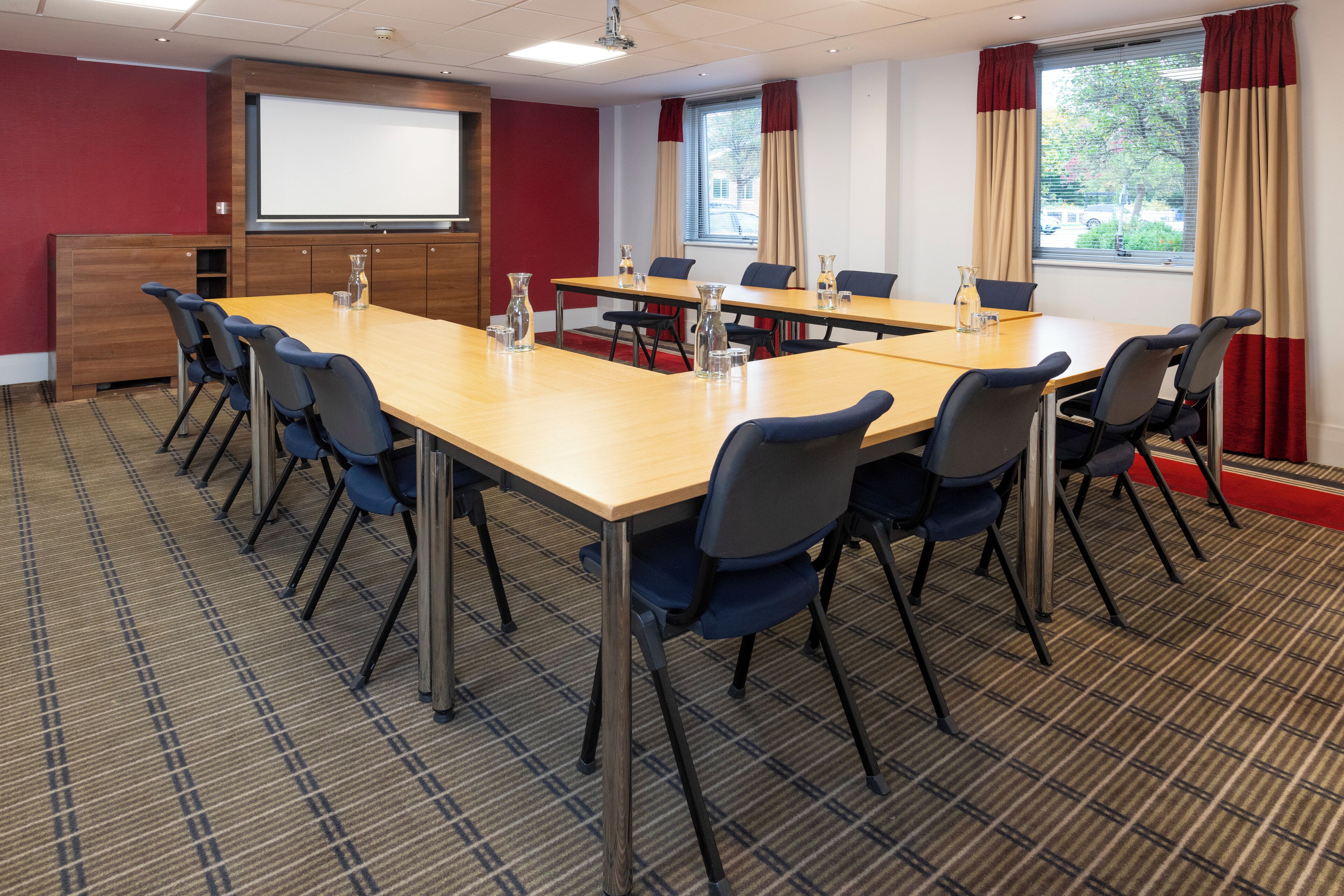 Un salón de conferencias con una gran mesa, sillas, una pantalla de proyector y una ventana con cortinas.
