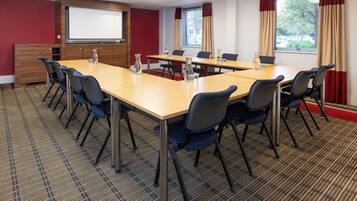 Un salón de conferencias con una gran mesa, sillas, una pantalla de proyector y una ventana con cortinas.