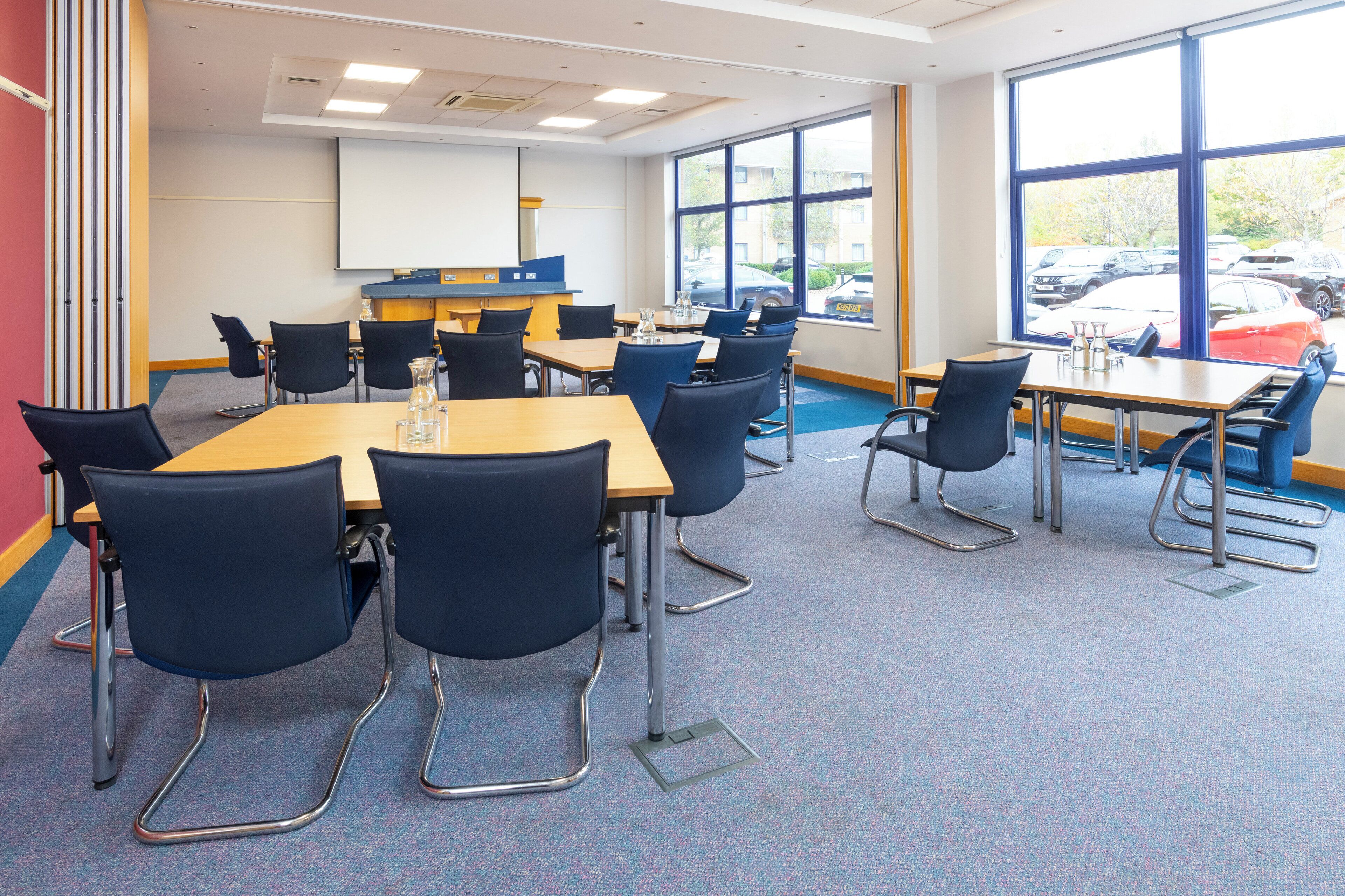 A conference room with a large table, chairs, a projector screen, and large windows with a parking view.