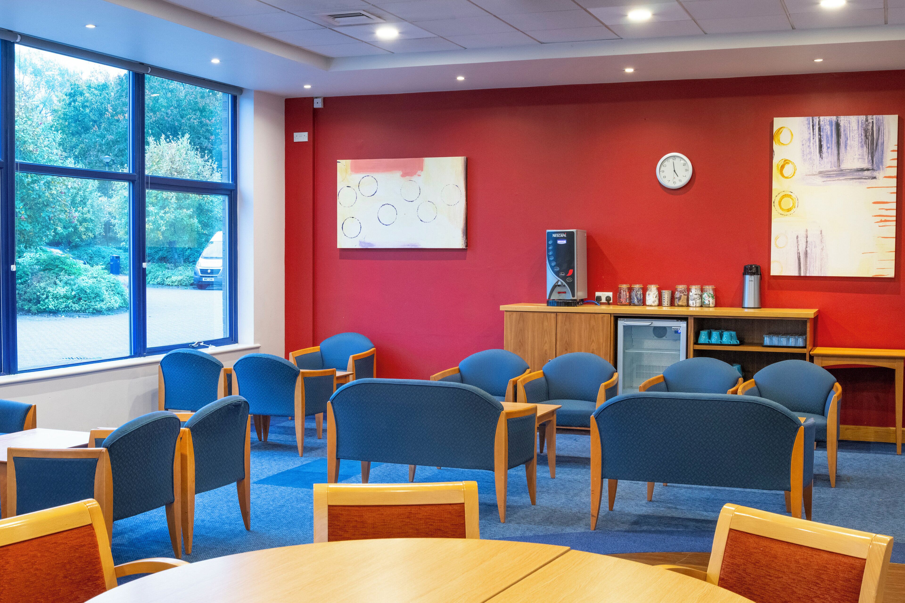 A library with blue chairs, wooden tables, a red wall, and a window with a view of trees.