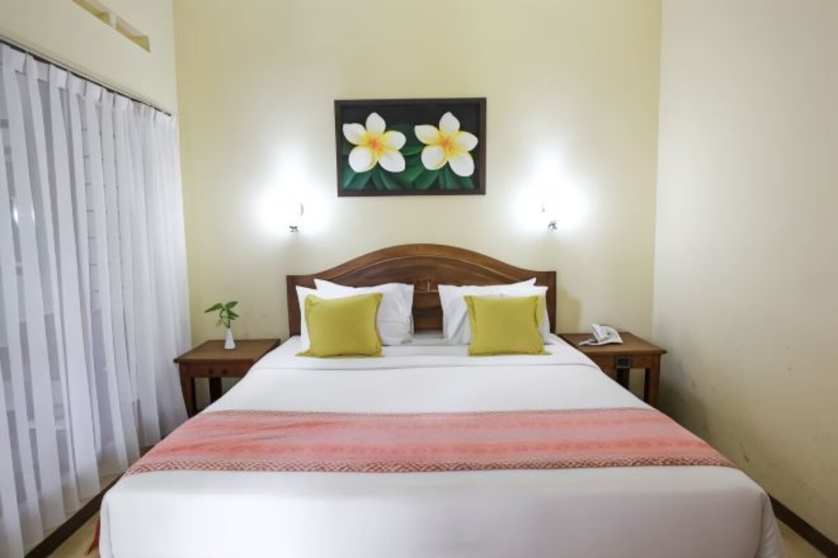 Ein Hotelzimmer mit Bett, zwei Nachttischen, einem Wandbild mit Blumen und einem Fenster mit Vorhängen.