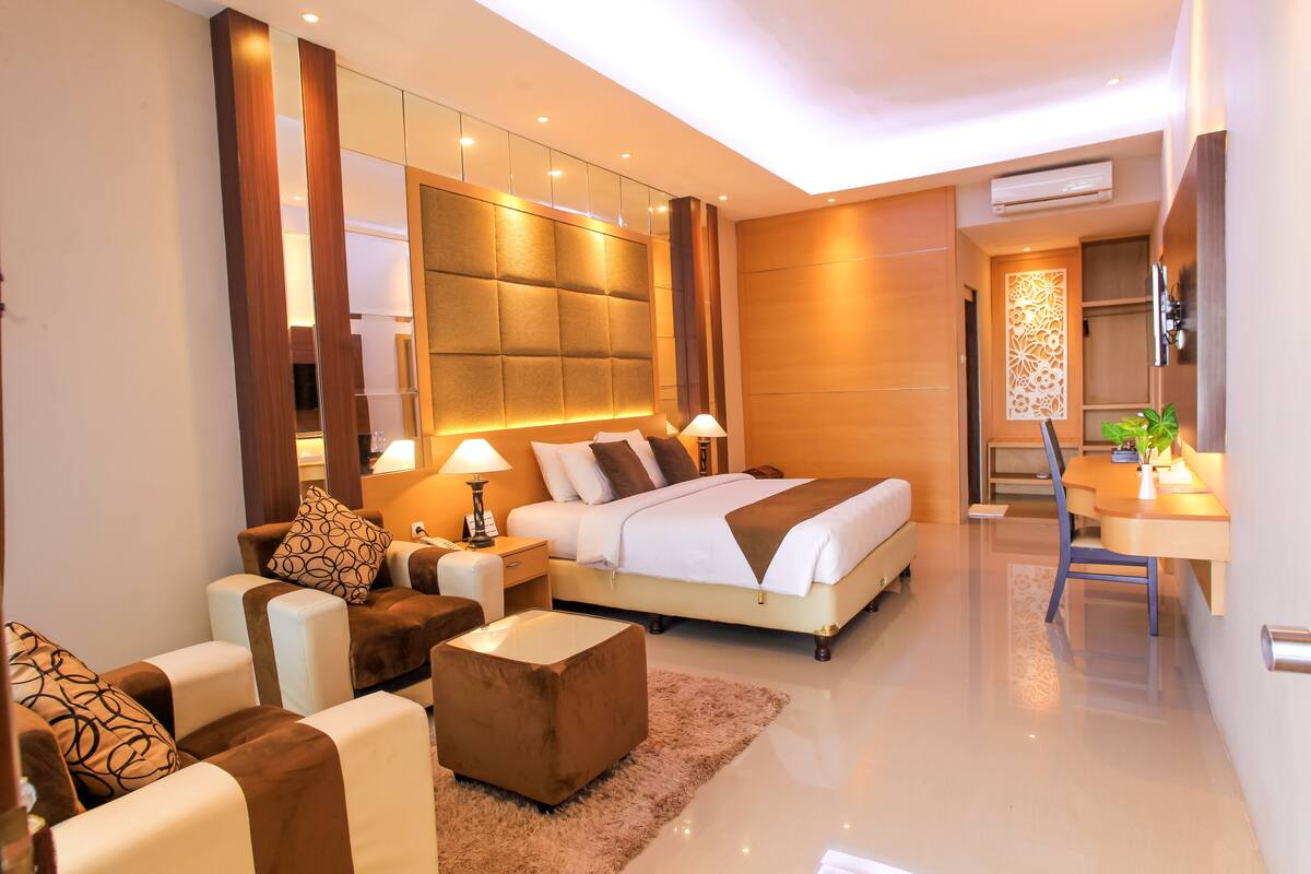 A modern hotel room with a large bed, a sitting area with a sofa and armchairs, a small table with a lamp, and a decorative wall panel.