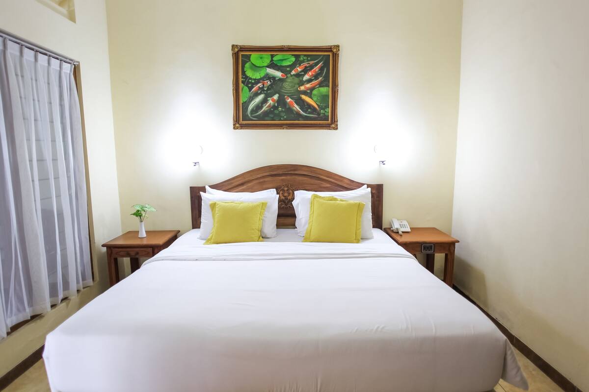 A neatly made bed with white sheets and two yellow pillows, a wooden headboard, a small bedside table with a plant, and a framed painting above the bed.