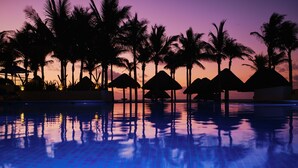 A pool with palm trees and thatched umbrellas in the background. - Hotel NYX Cancun - Near La Isla Shopping Mall (Cancun)