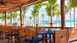 A covered outdoor seating area with wooden tables and chairs, overlooking a beach with palm trees and thatched umbrellas. - Hotel NYX Cancun - Near La Isla Shopping Mall (Cancun)