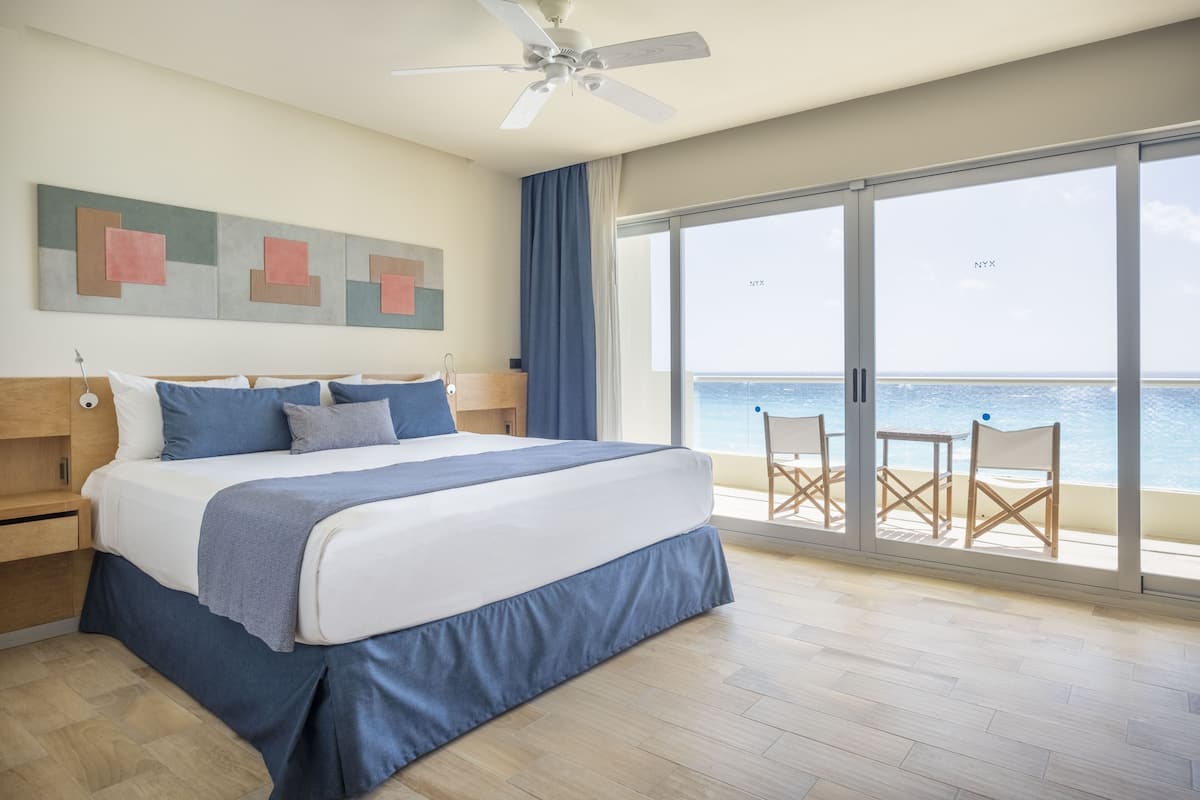 Un dormitorio con una cama grande, ventilador de techo, vistas al océano a través de ventanas amplias y un balcón con sillas.