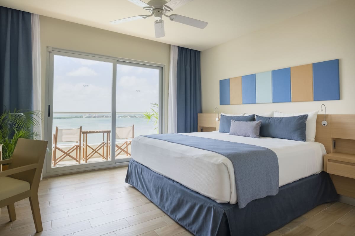 Habitación de hotel con cama, escritorio, silla, ventilador de techo, ventana grande con vista al mar, y una decoración en tonos azul y blanco.
