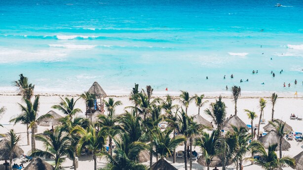 A beach with palm trees, thatched-roof huts, and people swimming in the water. - Hotel NYX Cancun - Near La Isla Shopping Mall (Cancun)