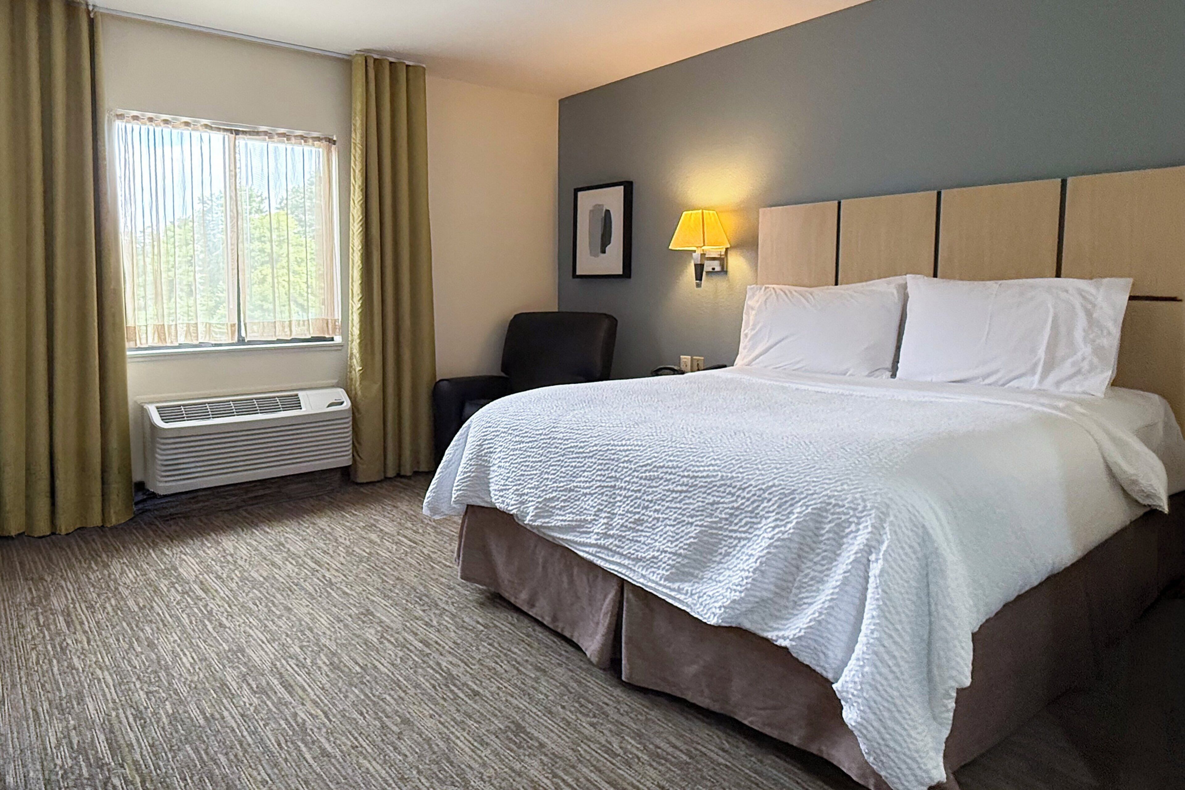 Habitación de hotel con una cama grande, una silla, una lámpara de pared y una ventana con cortinas.