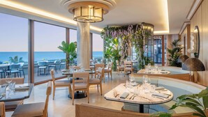 Un comedor moderno con mesas preparadas para dos, ventanas amplias que dan al mar y una columna central con vegetación.