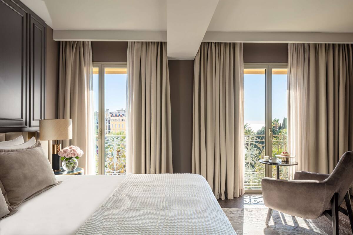 A hotel room with a large bed, a chair, a small table with a cup and flowers, and a balcony with curtains.