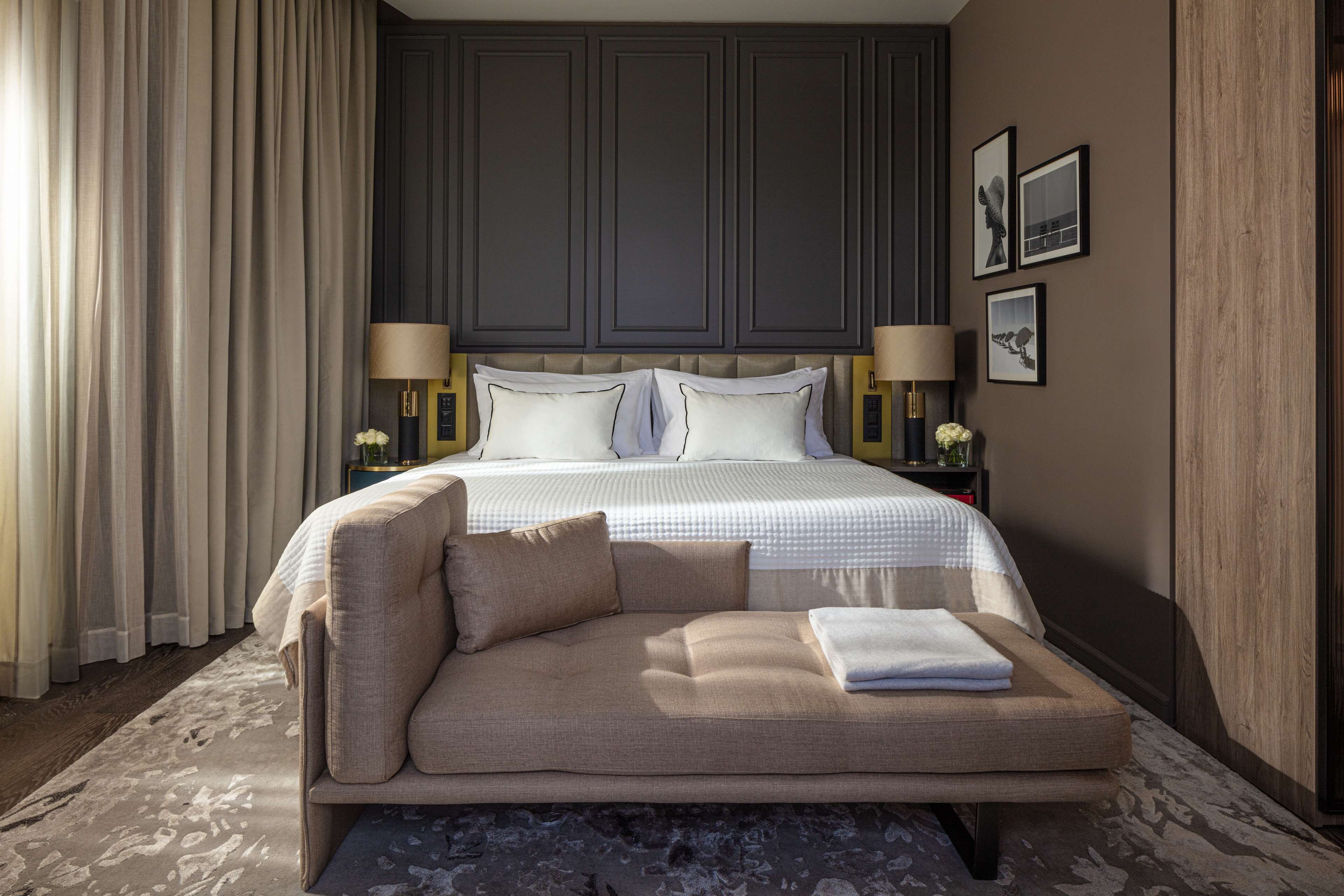 A bedroom with a bed, a sofa, a nightstand, lamps, and a wall with framed pictures.