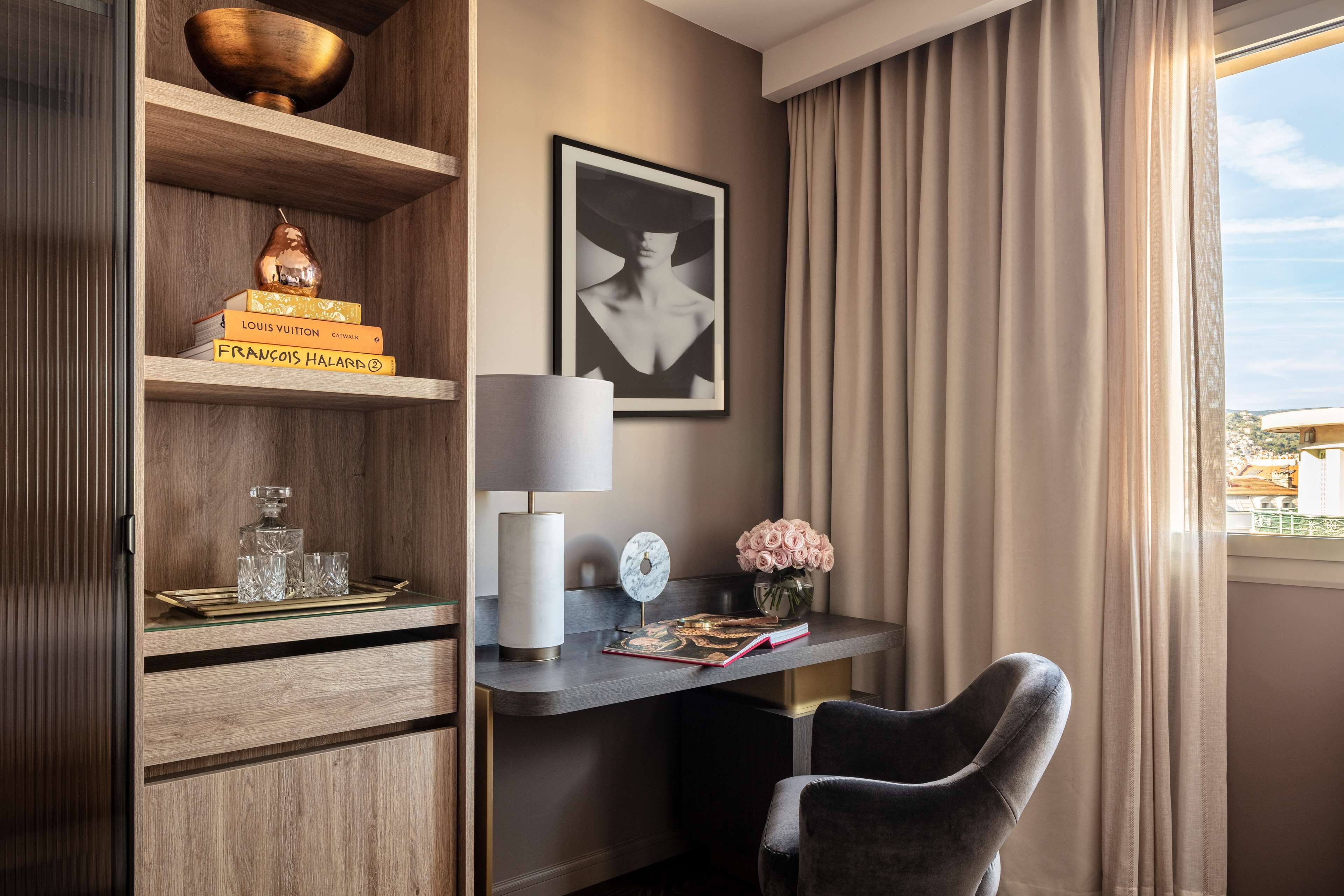 Una habitación moderna con un armario de madera, un escritorio con lámpara, una silla, una obra de arte enmarcada y vistas al exterior.