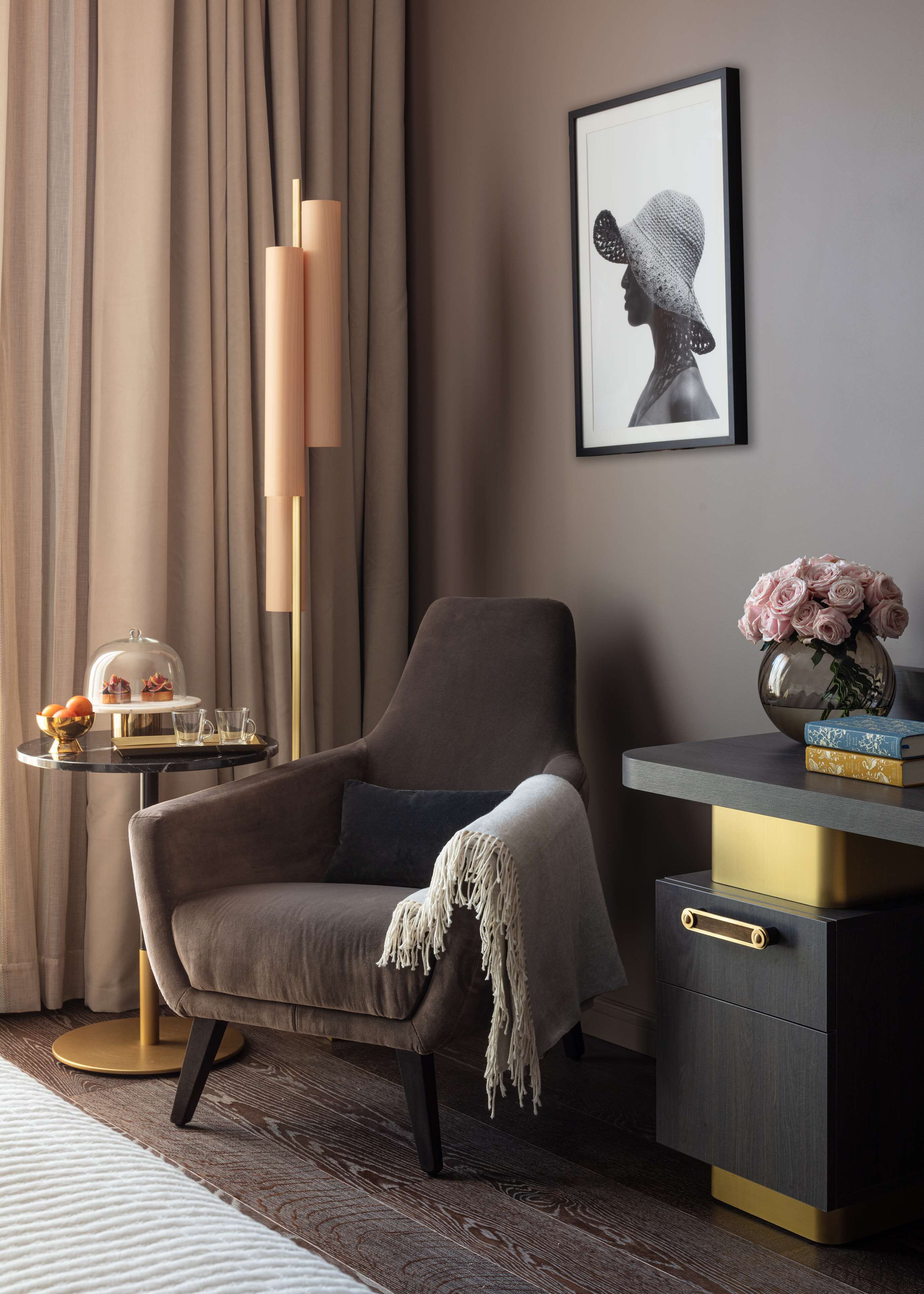 A modern room with a grey armchair, a side table with a glass dome, a tall floor lamp, a framed artwork, and a desk with a vase of flowers.