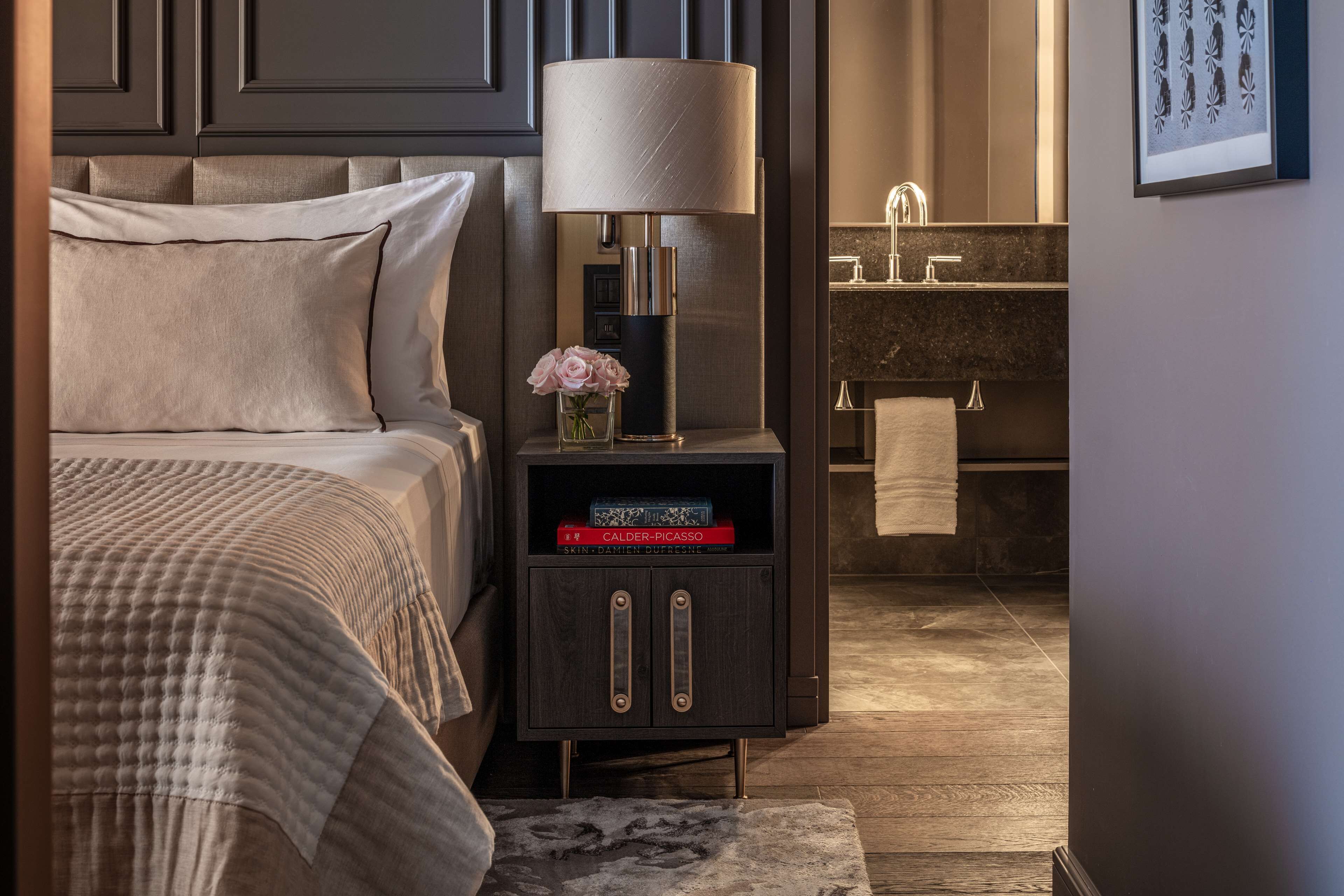 A bedroom with a bed, bedside table with a lamp, a vase with flowers, and a nightstand with books.