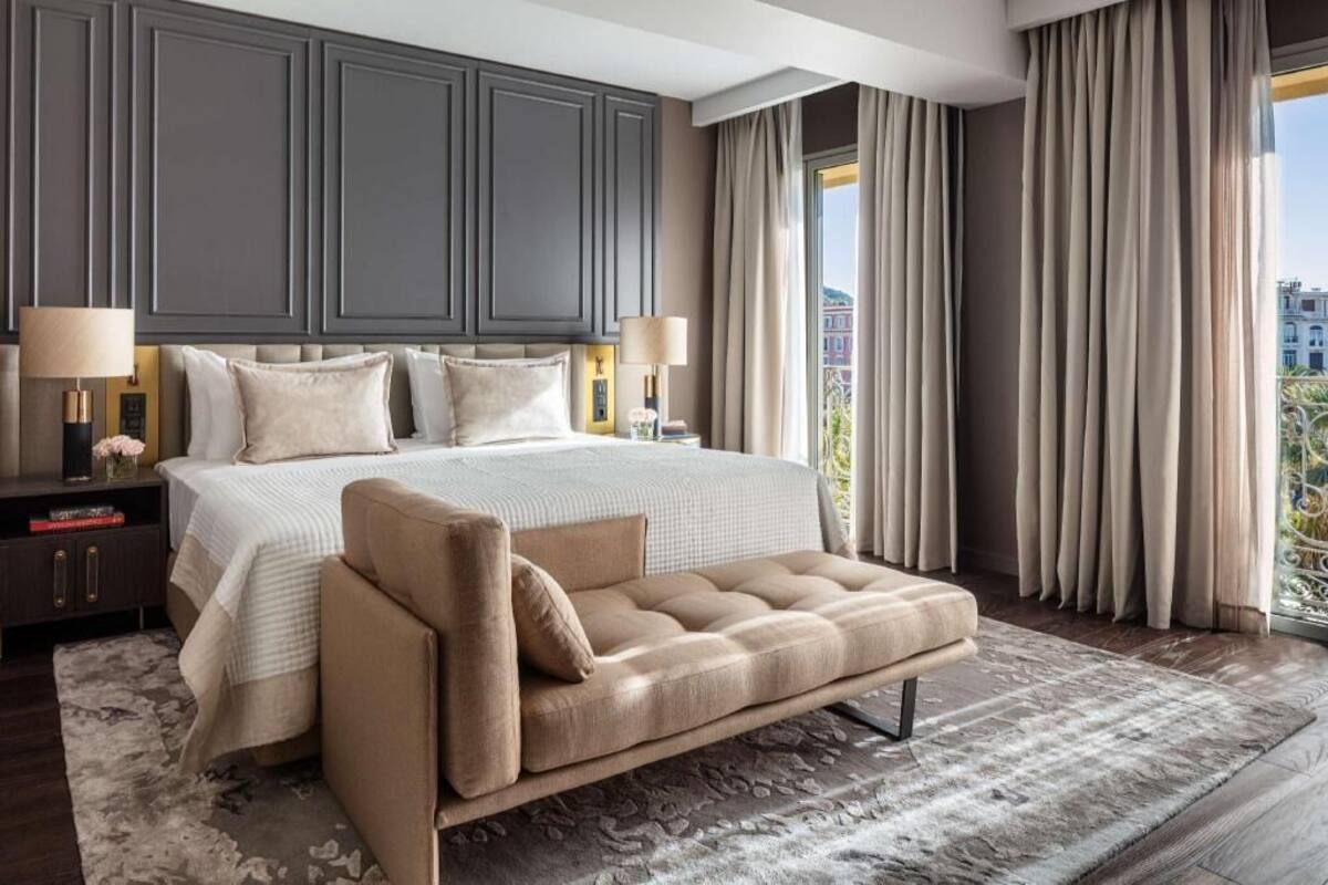 A bedroom with a large bed, a sofa, a nightstand, and a view of buildings through the window.