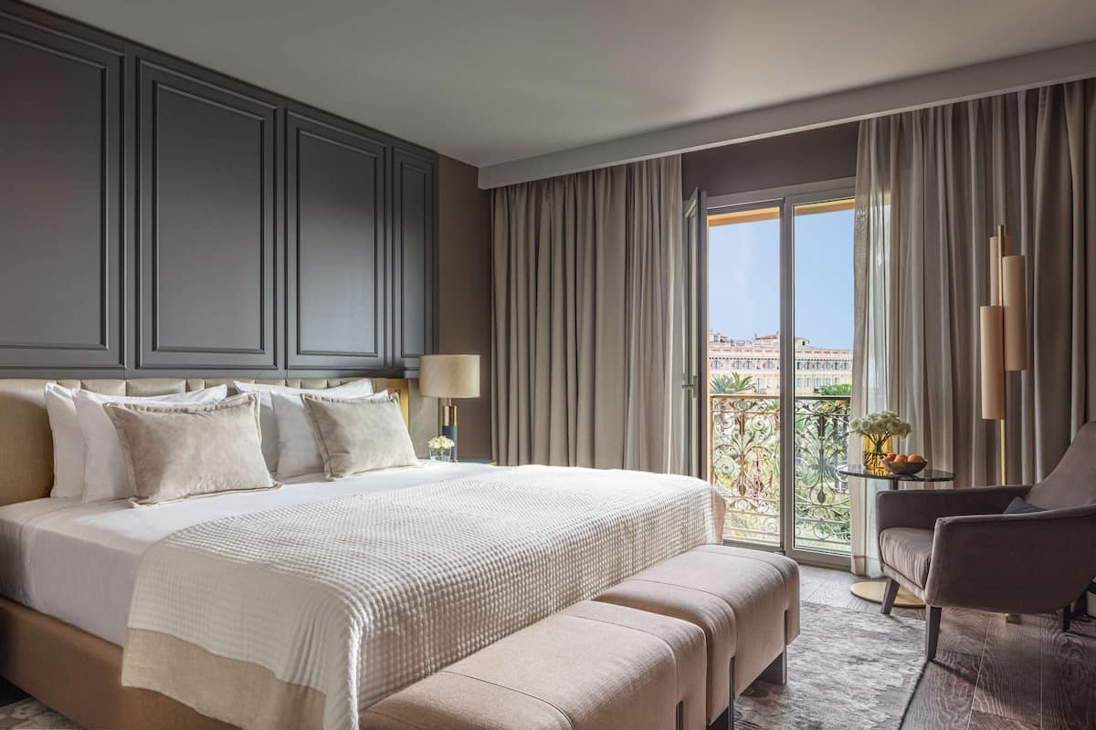 A hotel room with a large bed, a bedside table with a lamp, a chair, and a balcony with a view.