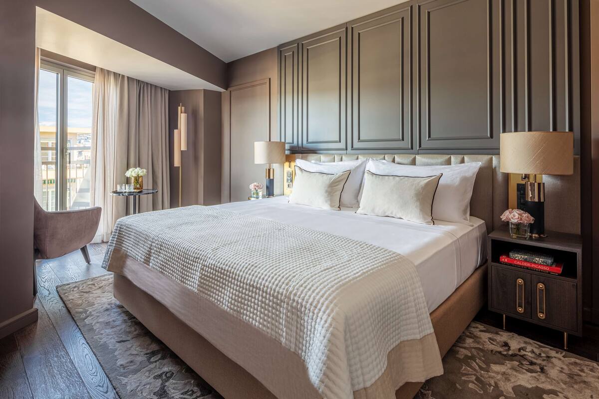 A hotel room with a large bed, a bedside table with a lamp, a chair, and a view of the city through the window.