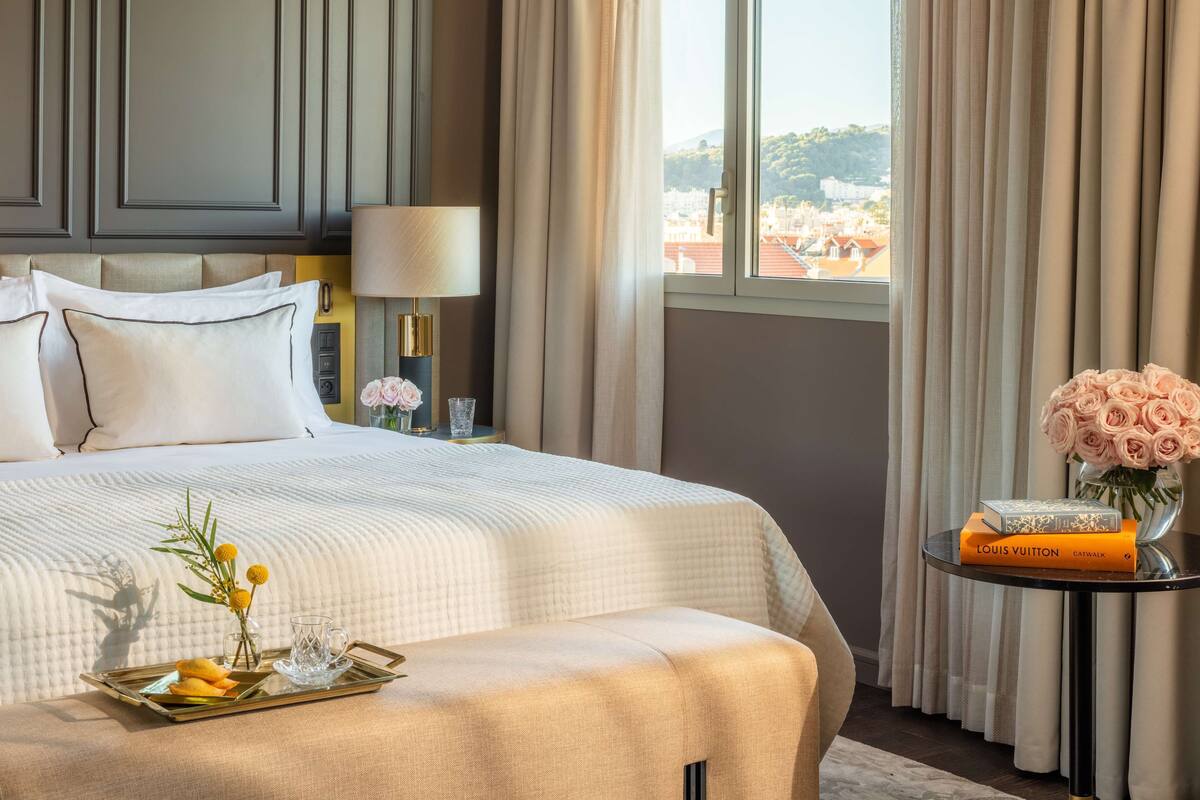 A hotel room with a bed, a bedside table with a lamp, a tray with food and flowers, and a view of buildings through the window.