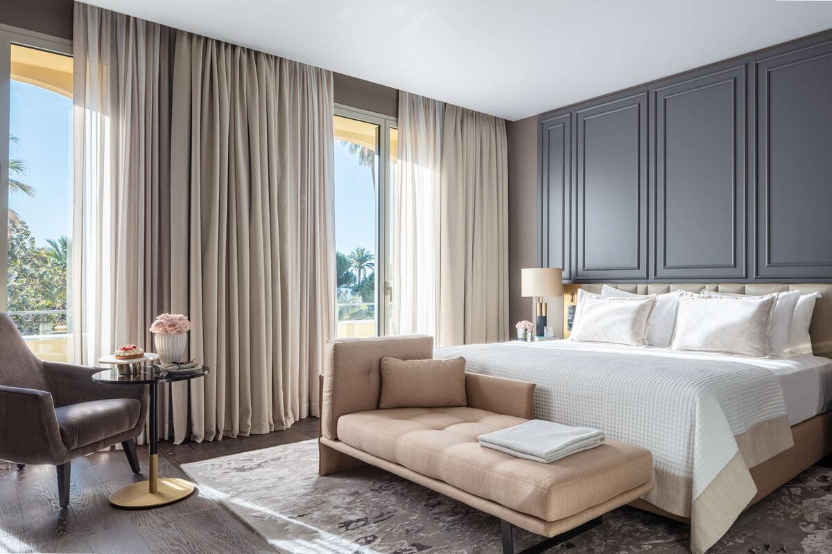 A modern hotel room with a large bed, a sofa, a small table with a vase of flowers, and a nightstand with a lamp.