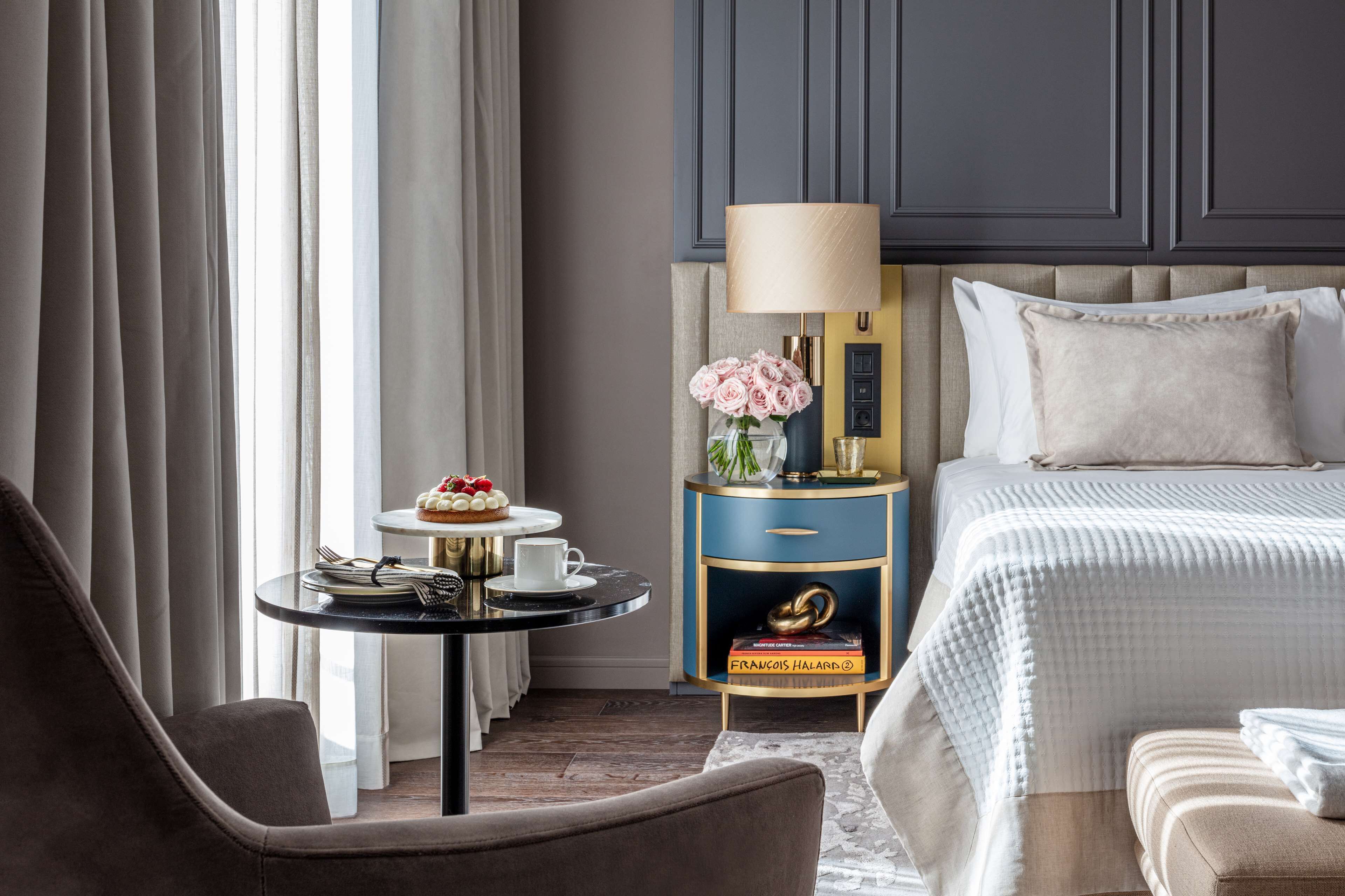 A hotel room with a bed, a round table with a cake, a lamp, and a nightstand with a book.