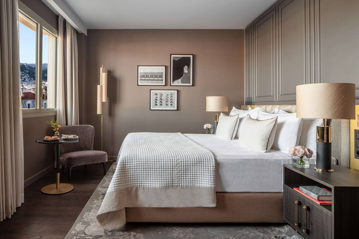 A hotel room with a bed, bedside lamps, a chair, a small table with a vase, and framed pictures on the wall.