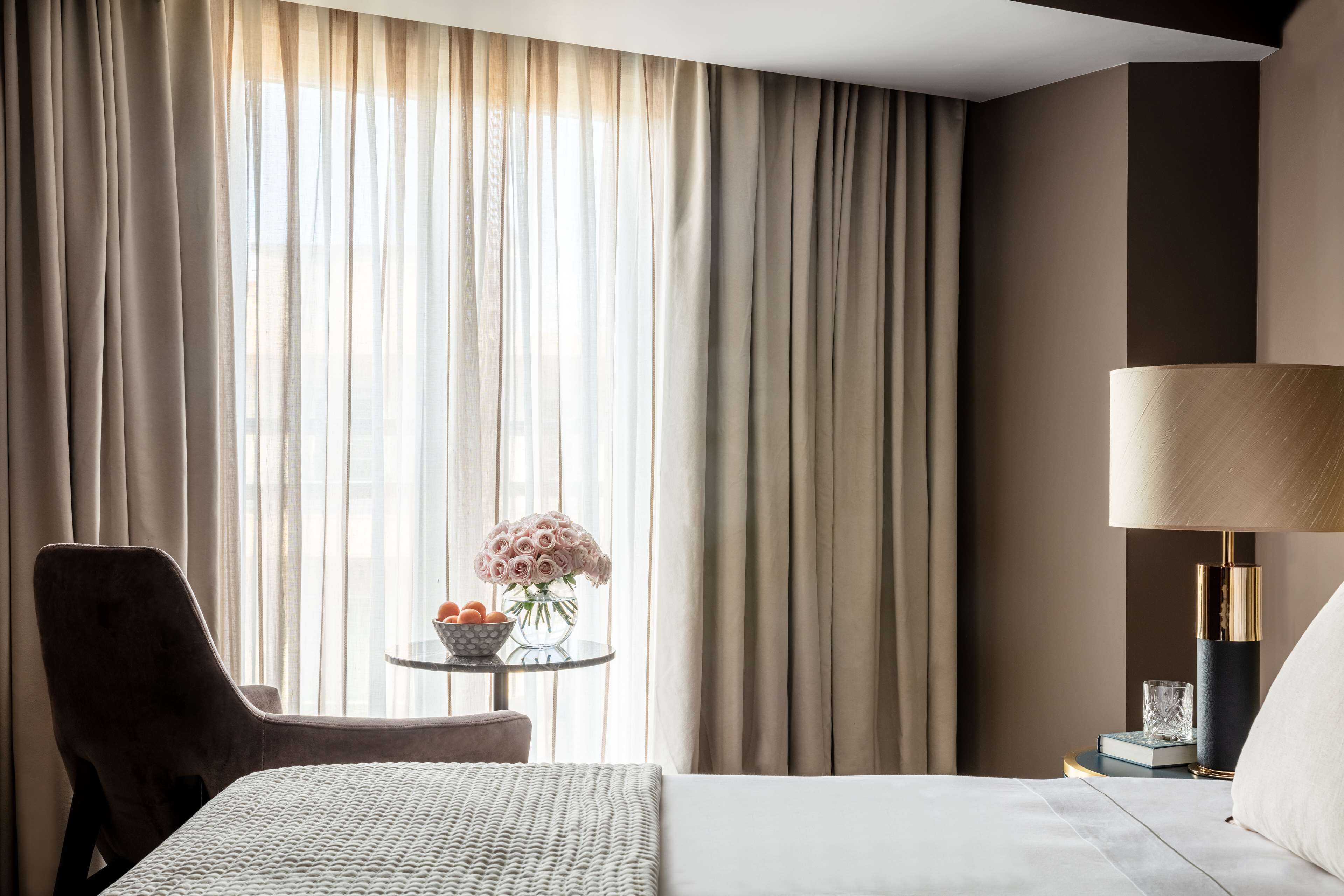 A hotel room with a bed, a chair, a small table with a vase of flowers, and a lamp.