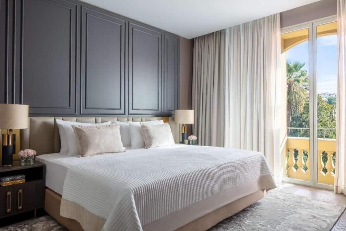 A bedroom with a large bed, two bedside tables with lamps, a balcony with curtains, and a view of greenery.