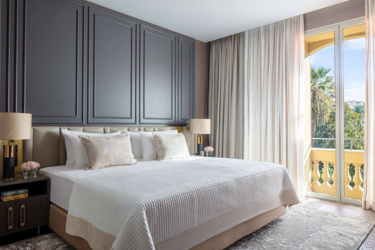 A large bed with a headboard, two bedside tables with lamps, and a view of a balcony with plants.