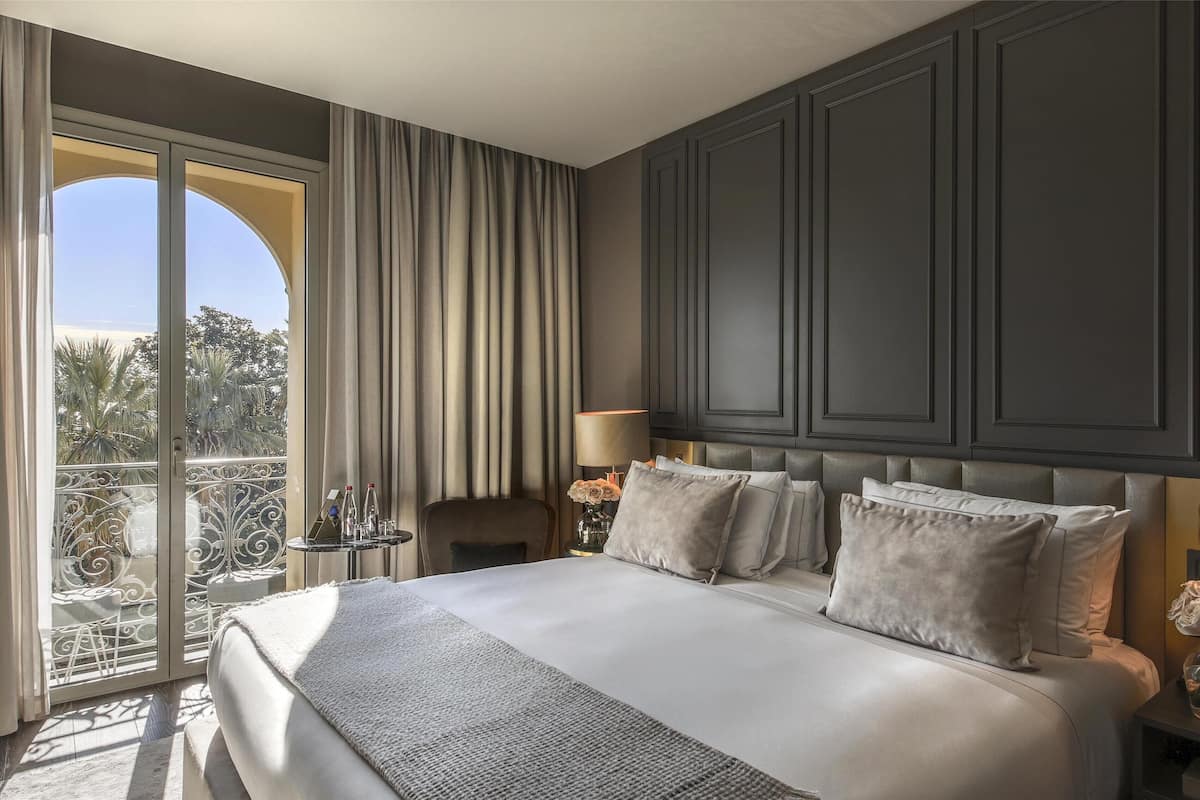 A hotel room with a large bed, two bedside tables, a view of palm trees, and a balcony.