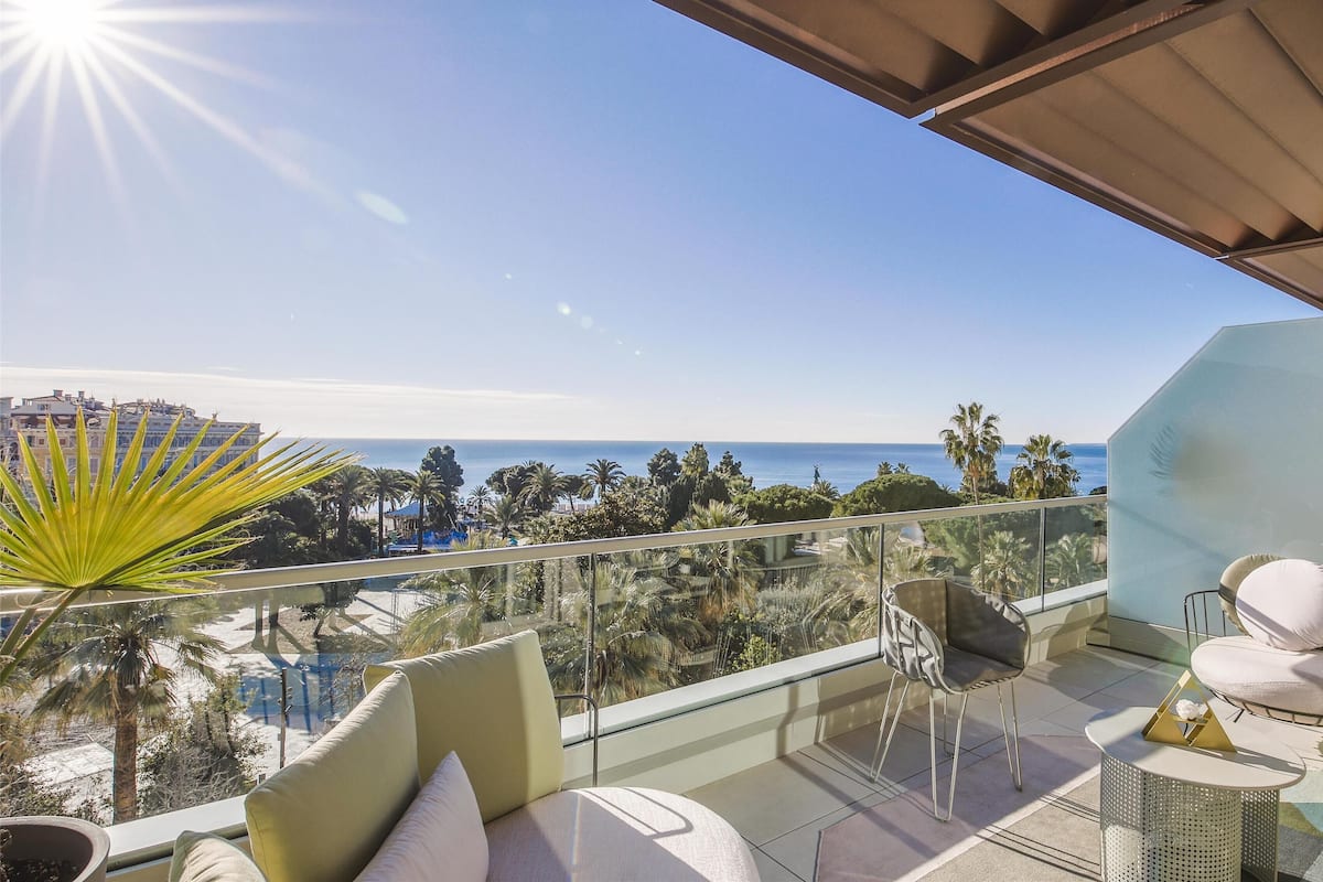 A balcony with a glass railing, outdoor furniture, and a view of the ocean and distant buildings.